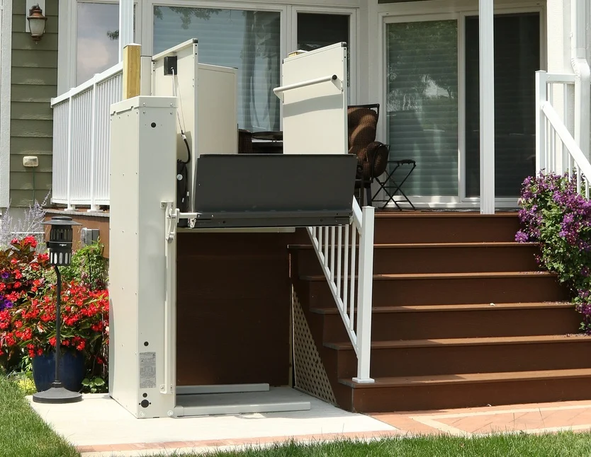 An outdoor vertical platform lift is installed next to a porch, providing wheelchair access alongside stairs, with flowers and patio furniture visible.