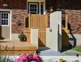 A vertical platform lift is installed next to wooden steps leading to a porch, providing wheelchair access to the brick house. Flowers and potted plants are in the foreground.