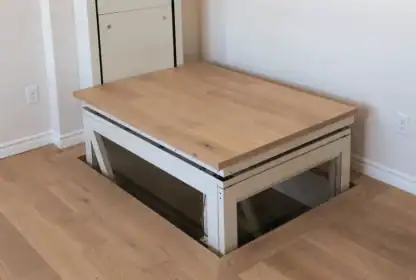 A section of wooden floor is raised by a mechanical platform, revealing glass sides and an opening below in a modern interior room.