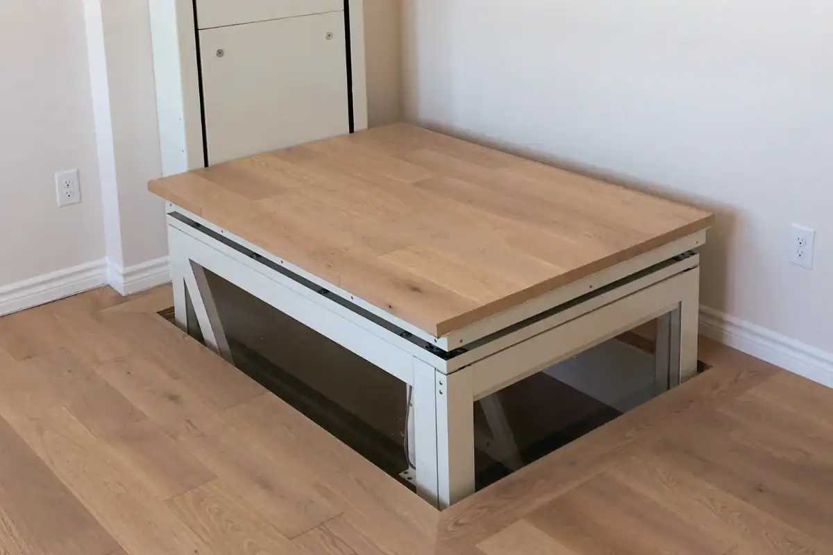 A section of wooden floor is raised by a mechanical platform, revealing glass sides and an opening below in a modern interior room.