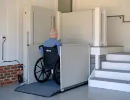 An older adult in a wheelchair uses a vertical lift to move from the garage floor to the elevated indoor area next to a staircase.
