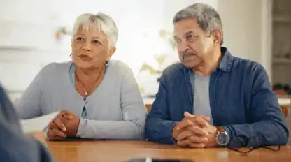 An older woman and man sit side by side at a table, looking attentively toward someone off camera, with their hands folded on the tabletop.