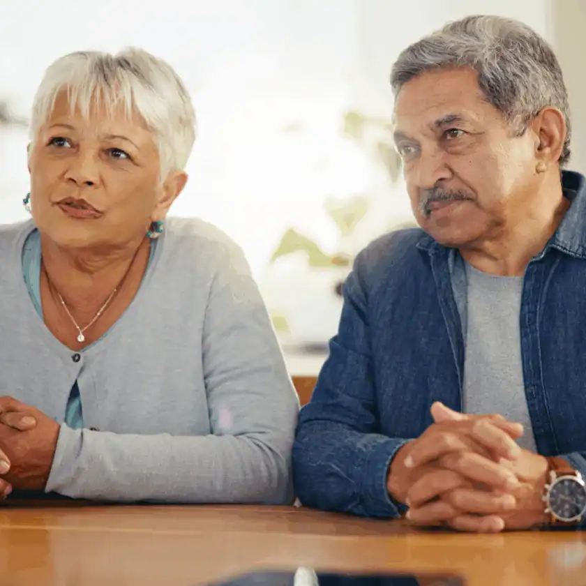 An older woman and man sit side by side at a table, looking attentively toward someone off camera, with their hands folded on the tabletop.