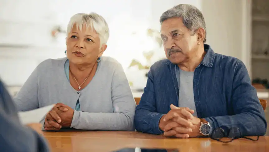 An older woman and man sit side by side at a table, looking attentively toward someone off camera, with their hands folded on the tabletop.