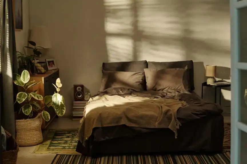 A neatly made bed with dark bedding in a cozy, sunlit bedroom; a plant, lamp, and books are visible beside the bed.