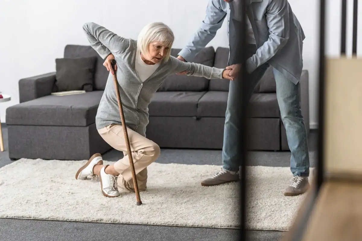 An elderly woman with a cane is assisted by a person as she struggles to get up from a kneeling position on a carpeted floor in a living room.