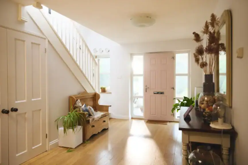 A bright hallway with wooden floors, a pink front door, staircase, wooden bench with cushions, potted plants, and a console table with decorative items.