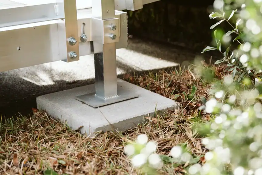 A metal post supports a structure, anchored to a concrete slab base on the ground surrounded by grass and small plants.