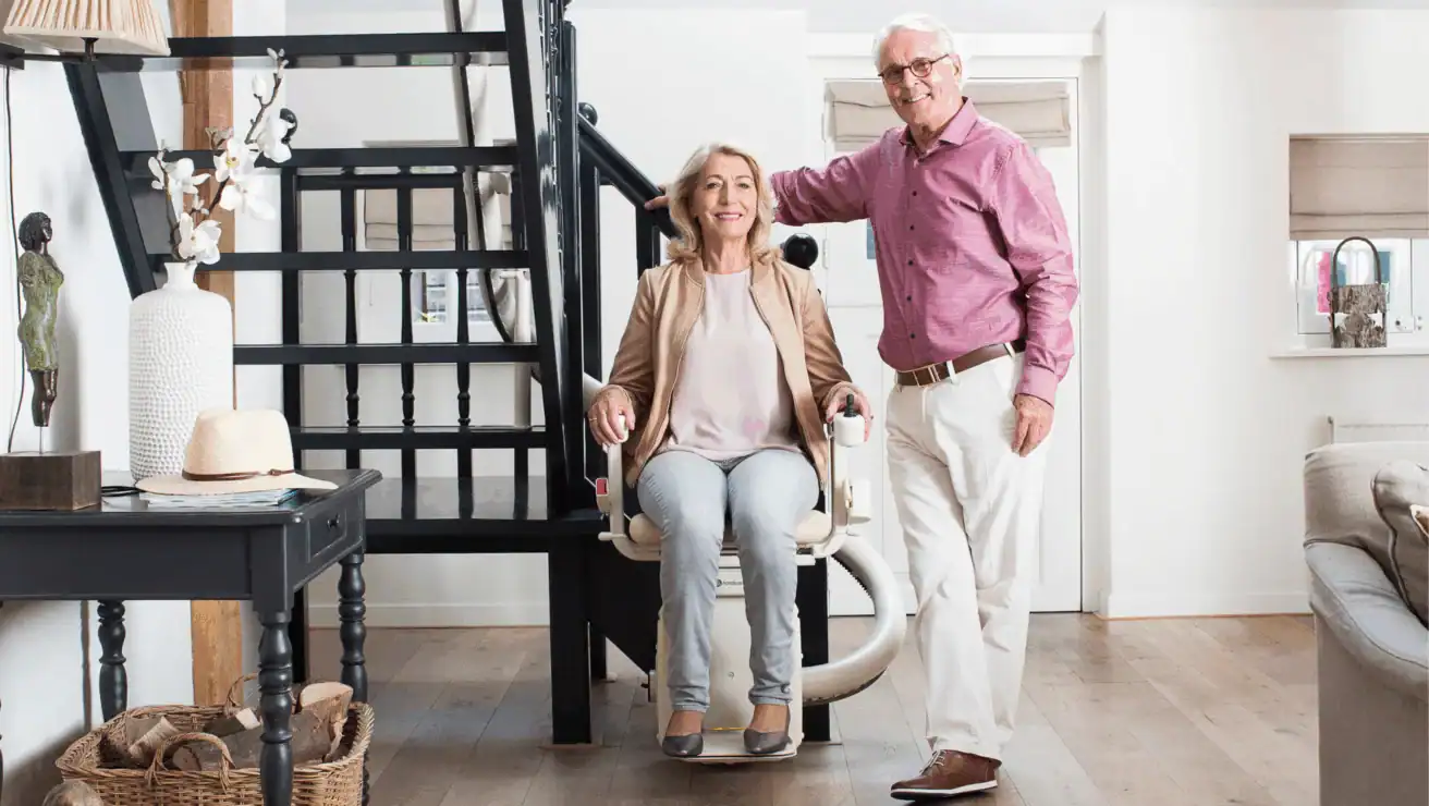 An older woman sits on a stairlift at the base of a staircase, smiling, while an older man stands beside her with his hand on her shoulder in a modern living room.