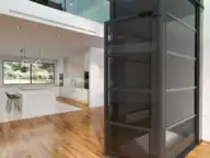 A modern glass residential elevator in a spacious open-plan living area with wood floors, white walls, and a kitchen visible in the background.