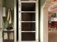 A modern residential elevator with glass doors is installed in a hallway next to a coat rack, bench, and mirror, leading into a dining area.
