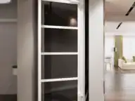 A modern residential elevator with a glass door is situated between a hallway with marble flooring and a living room with wooden floors and large windows.