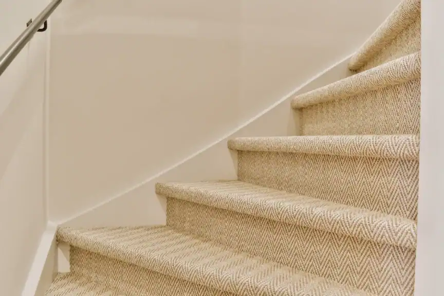 A close-up view of carpeted stairs with a light herringbone pattern and a silver handrail on the left wall.