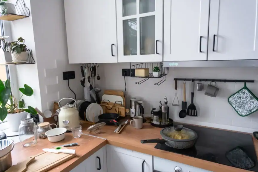 A kitchen with white cabinets, various utensils, and dishes on the countertop and racks; a pot is cooking on the stove and plants are by the window.