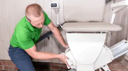 A man in a green shirt wipes down a beige stair lift chair with a cloth, cleaning its base near a staircase.