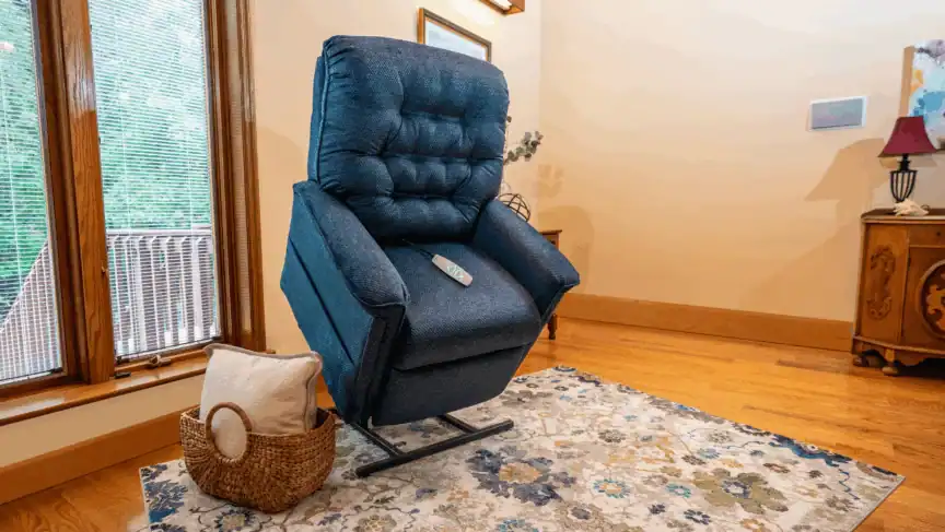 A blue upholstered lift chair is partially elevated in a living room with a floral rug, wooden floors, and a basket with a pillow beside it. A remote control rests on the chair seat.