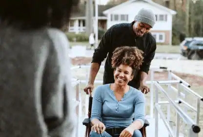 A smiling woman in a wheelchair is being pushed up a ramp by a man; another person stands in the foreground. A house is visible in the background.