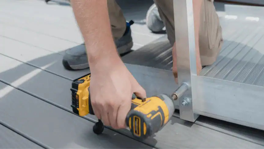 A person uses a yellow cordless power drill to fasten a bolt into a metal frame on a gray deck.