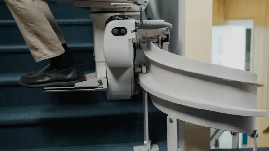 A person riding a stairlift chair up a carpeted staircase, with only their legs and the stairlift mechanism visible.
