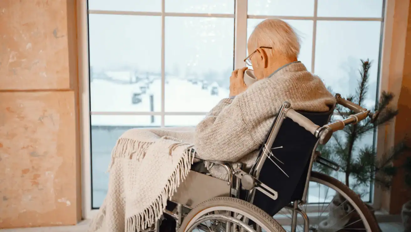 An elderly person in a wheelchair, wrapped in a blanket and shawl, drinks from a cup while looking out a large window at a snowy landscape.