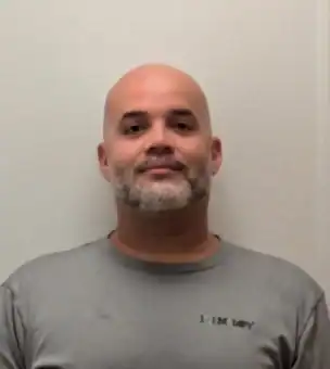 A man with a shaved head and short beard is standing against a plain light-colored wall, wearing a gray t-shirt with a small logo on the left chest.