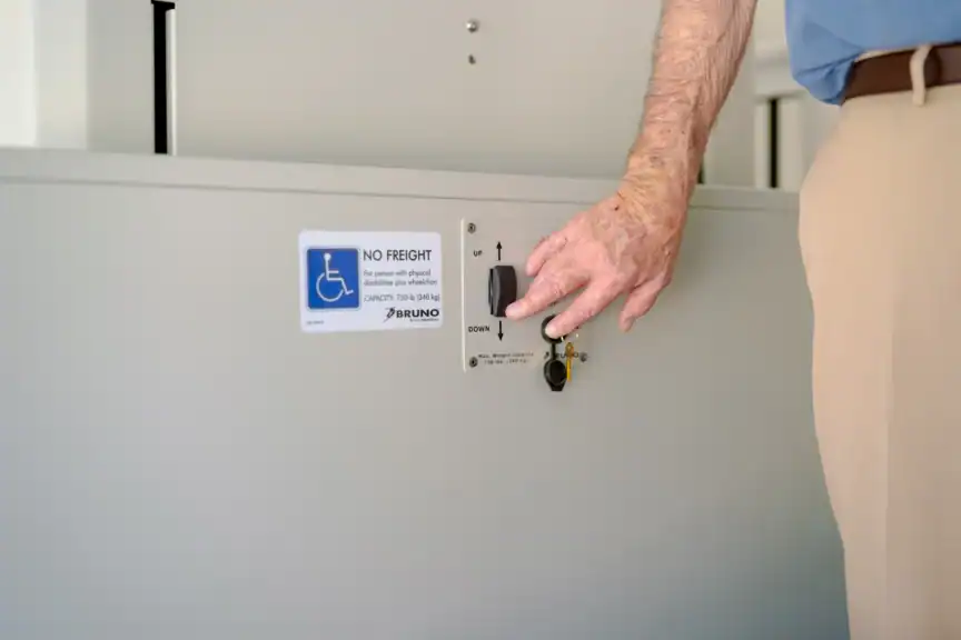 A person’s hand presses the control switch on a wheelchair lift panel labeled with accessibility and operation instructions.
