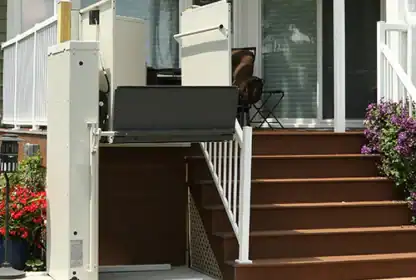 A vertical platform lift is installed next to stairs leading to a porch, providing wheelchair access to the elevated entrance of a house.