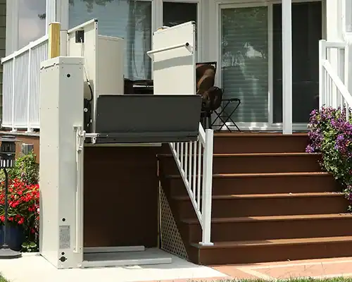 A vertical platform lift is installed next to stairs leading to a porch, providing wheelchair access to the elevated entrance of a house.