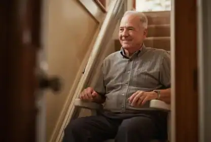 An older man sits on a stairlift at the bottom of a carpeted staircase, smiling in a well-lit home corridor.