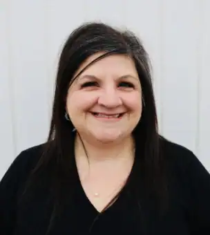 A woman with long dark hair and a black top stands smiling in front of a light-colored wooden wall.