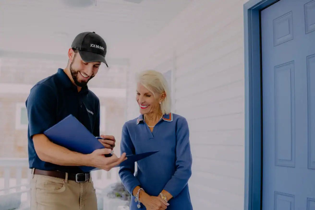 A man in a uniform shows documents to an older woman on a porch next to a blue door. Both are smiling and engaged in conversation.