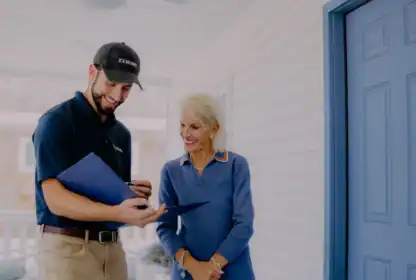 A man in a uniform shows documents to an older woman on a porch next to a blue door. Both are smiling and engaged in conversation.
