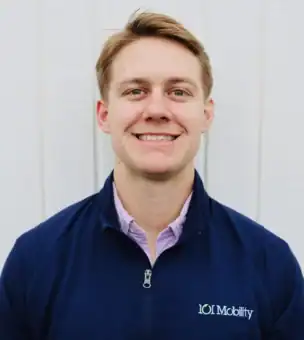 A man wearing a dark blue 101 Mobility jacket and a light purple shirt stands in front of a white wooden wall, smiling at the camera.