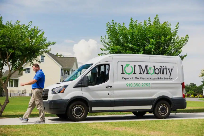 A man walks next to a white 101 Mobility van parked on a suburban street, with trees and a house in the background.