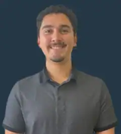 A man with short dark hair and a mustache smiles at the camera, wearing a dark short-sleeve collared shirt, standing in front of a plain dark background.