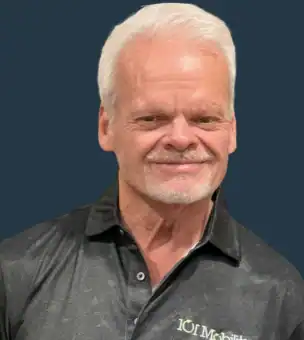 Man with white hair and goatee wearing a black collared shirt with "IOT Mobility" logo, posing against a solid dark blue background.