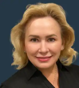 A woman with blonde hair and light skin smiles at the camera, wearing a black top and small earrings, against a dark blue background.