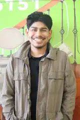 A man with dark hair and a beard smiles at the camera, wearing a brown jacket over a black shirt, standing in front of a green and orange background.