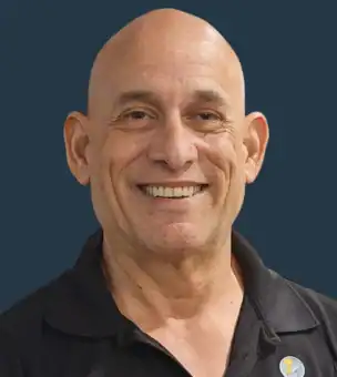 A smiling bald man wearing a black collared shirt and a round pin, posed in front of a plain dark background.