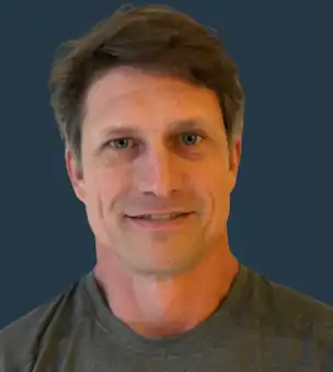 A middle-aged man with short brown hair and a slight smile wearing a gray shirt, pictured against a solid dark blue background.