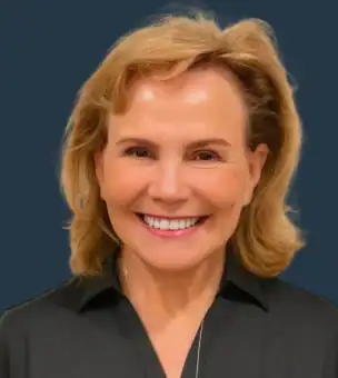 A middle-aged woman with blonde hair, wearing a black collared shirt, smiles at the camera against a plain dark blue background.