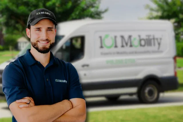 A man in an IOT Mobility uniform stands with arms crossed in front of a white IOT Mobility van parked outdoors.
