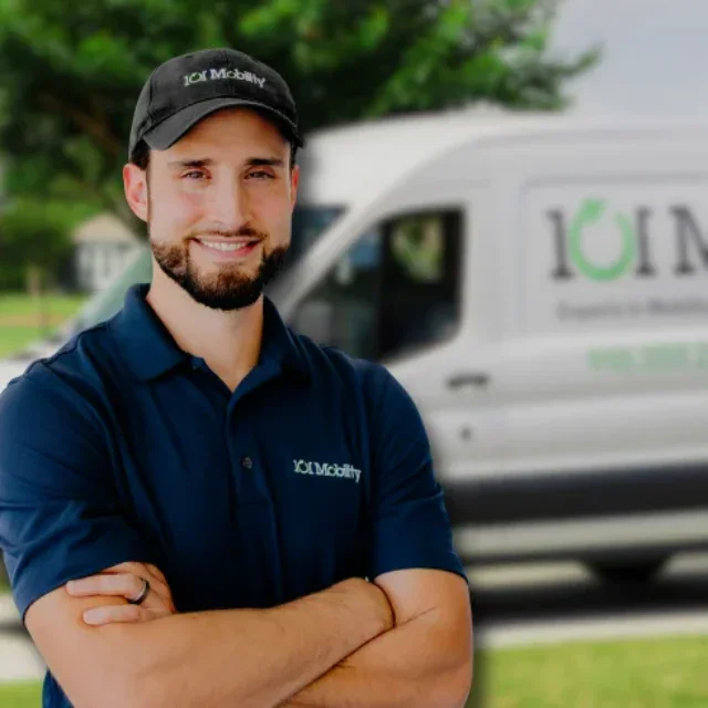 A man in an IOT Mobility uniform stands with arms crossed in front of a white IOT Mobility van parked outdoors.