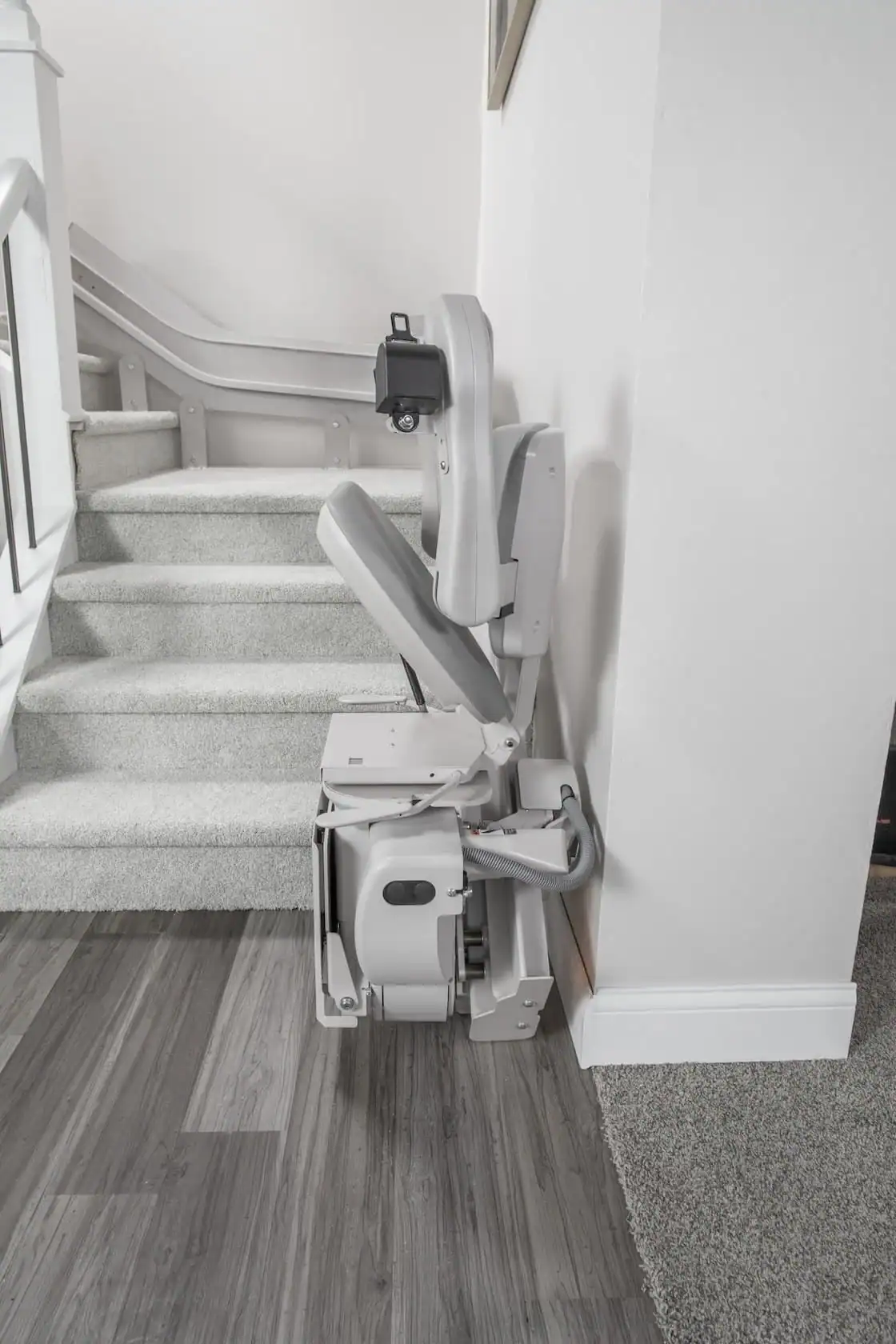 A stair lift installed along a carpeted staircase, with the chair folded up against the wall at the base of the stairs.