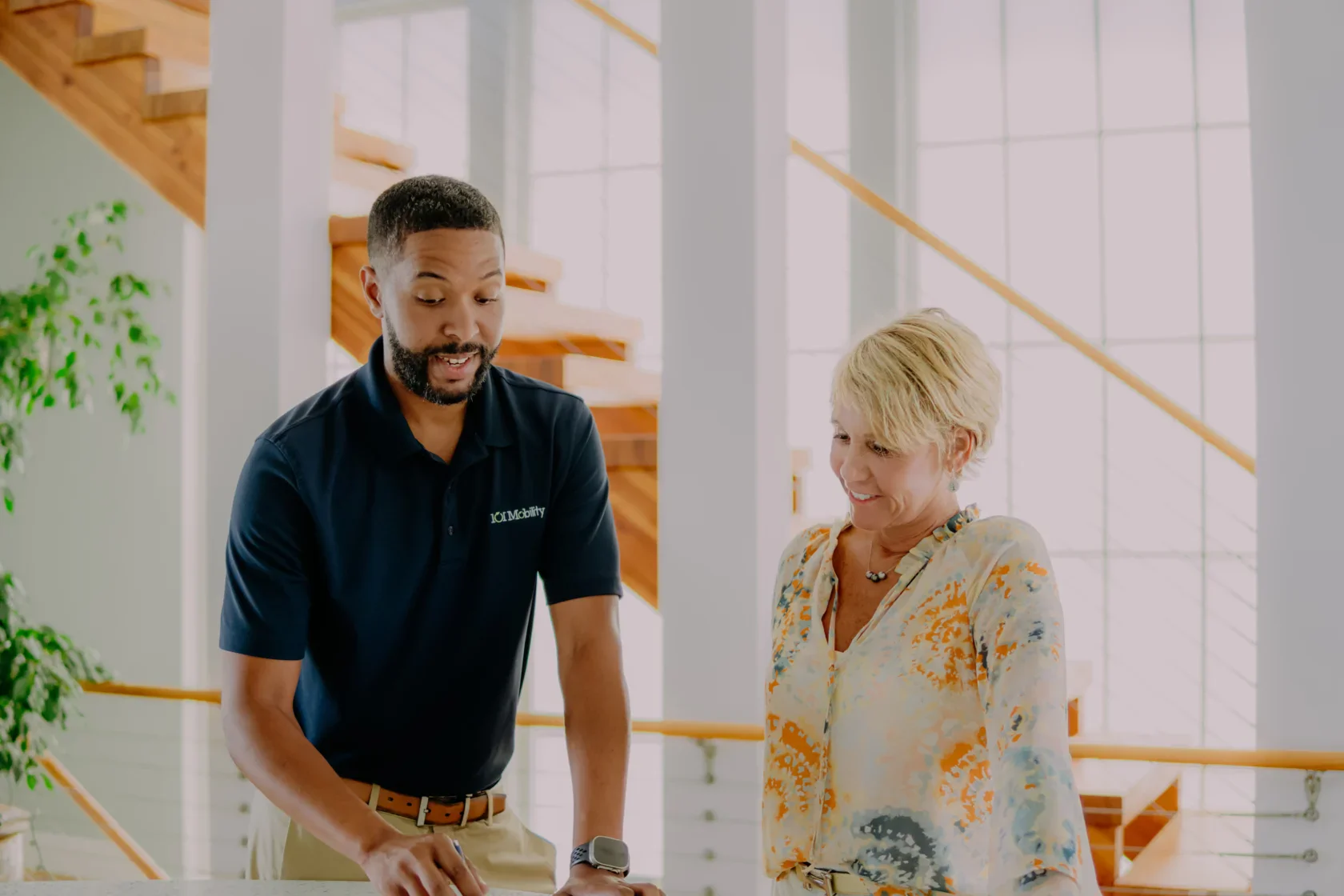 Two people stand indoors by a staircase, looking at something on a table. The man is talking and gesturing, while the woman stands beside him, watching and smiling.