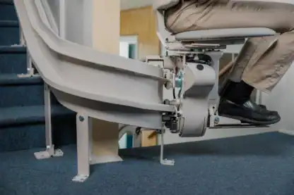 Close-up of a stairlift carrying a seated person up a carpeted staircase, showing the mechanical track and part of the person's legs and feet.