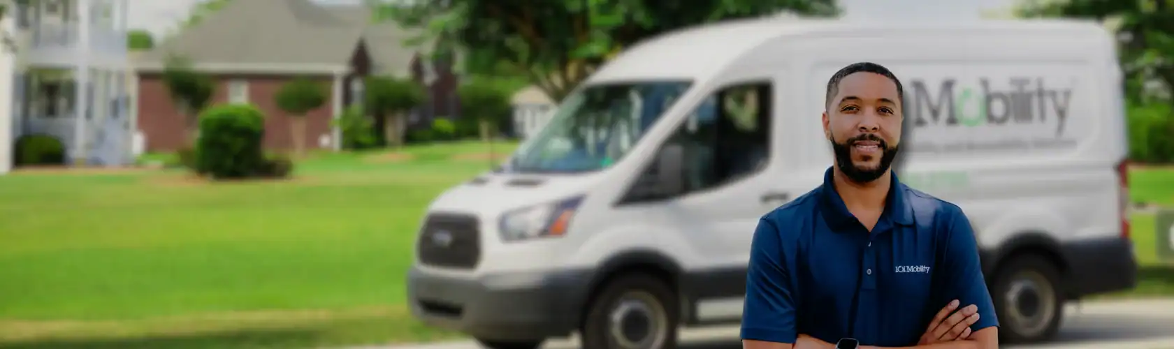 A man in a blue polo shirt stands outside with his arms crossed in front of a white van parked on a residential street.