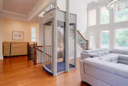 A modern residential elevator is installed in a spacious living room with hardwood floors, large windows, and a light gray sectional sofa.
