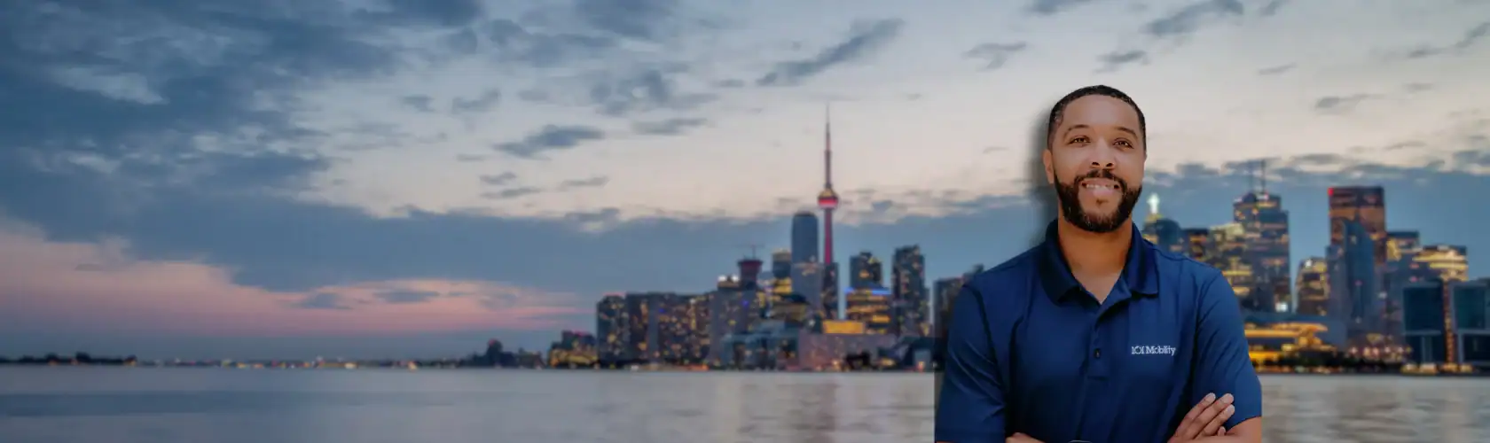Man in a blue polo shirt standing in front of a waterfront city skyline at dusk, with buildings and a prominent tower in the background.