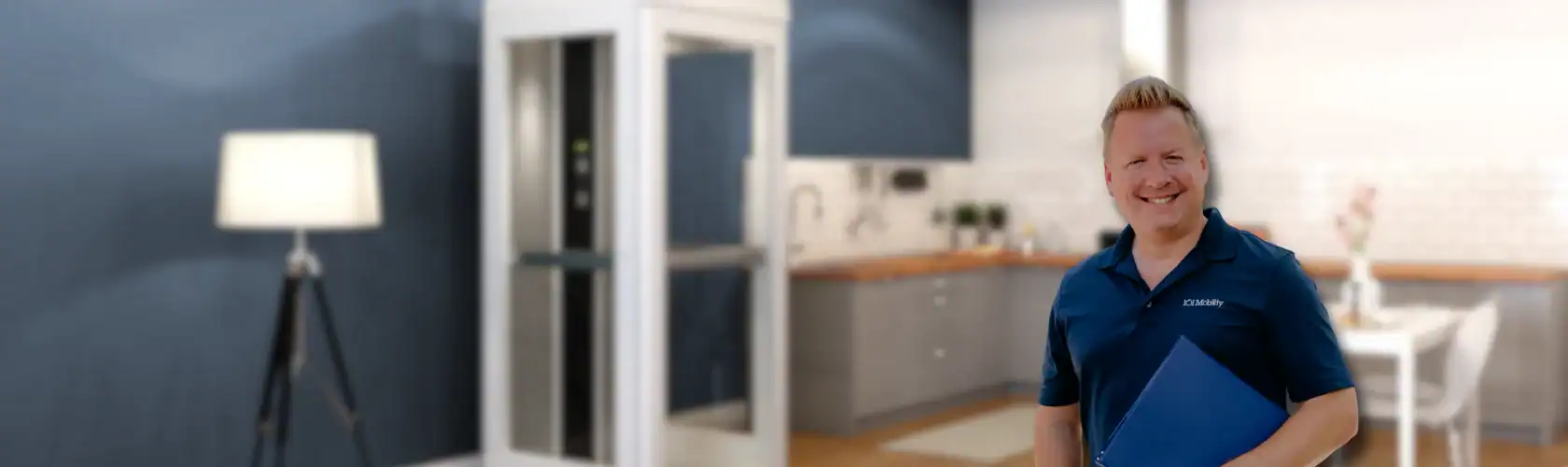 A man in a blue shirt holding a folder stands smiling in a modern kitchen and living area with an elevator in the background.
